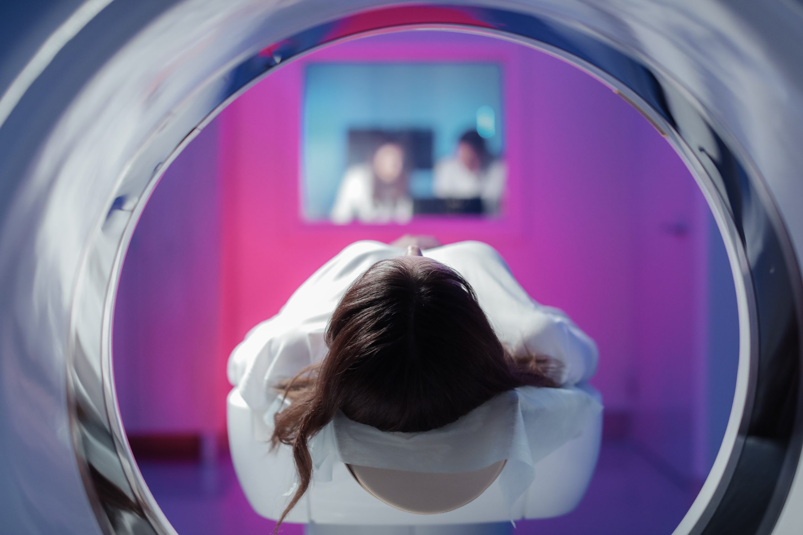 The girl patient is lying in the tomograph and waiting for a scan. Three doctors from the exam room look at the pictures